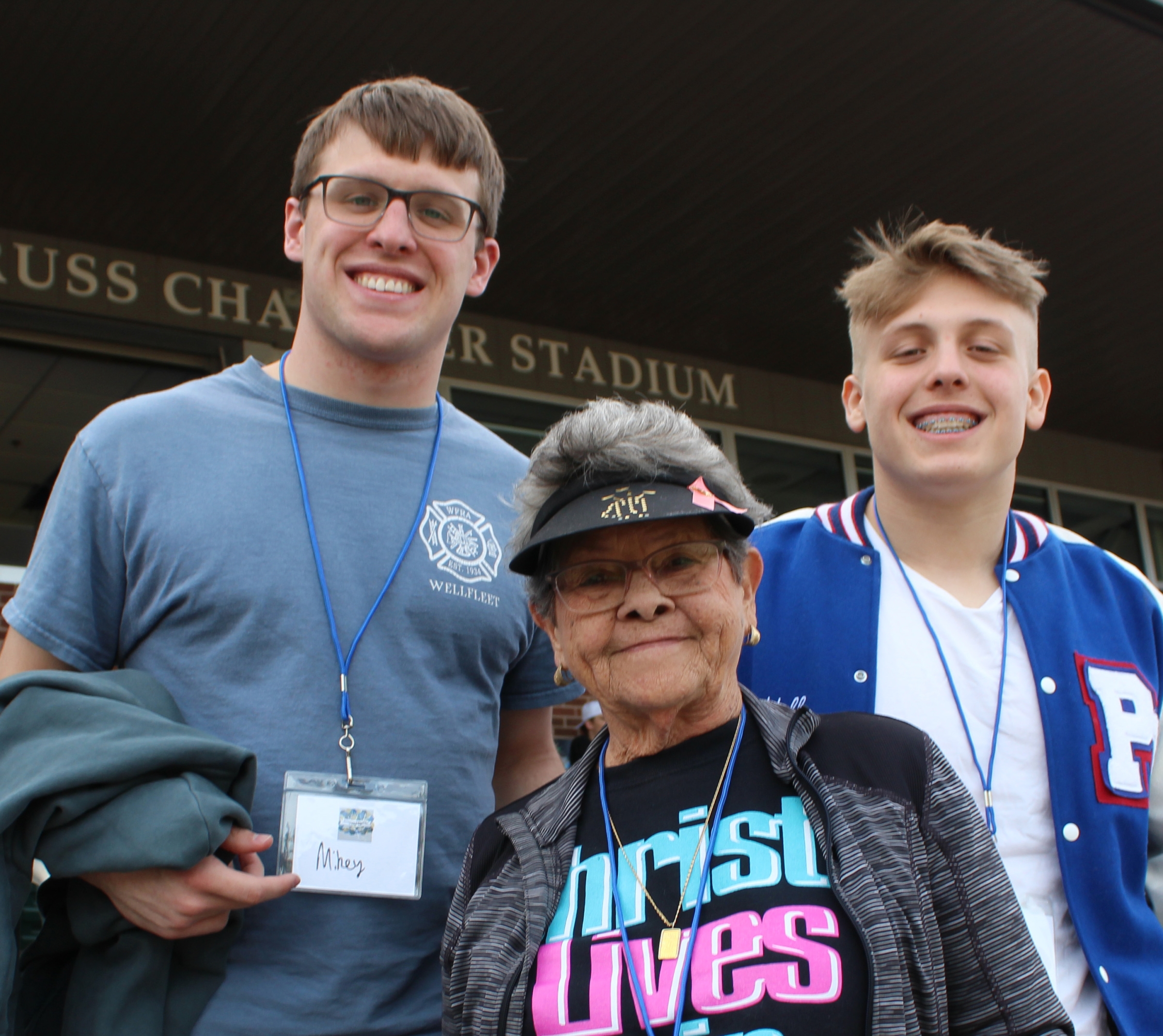 Two young men, smiling, stand on either side of an older woman wearing a visor