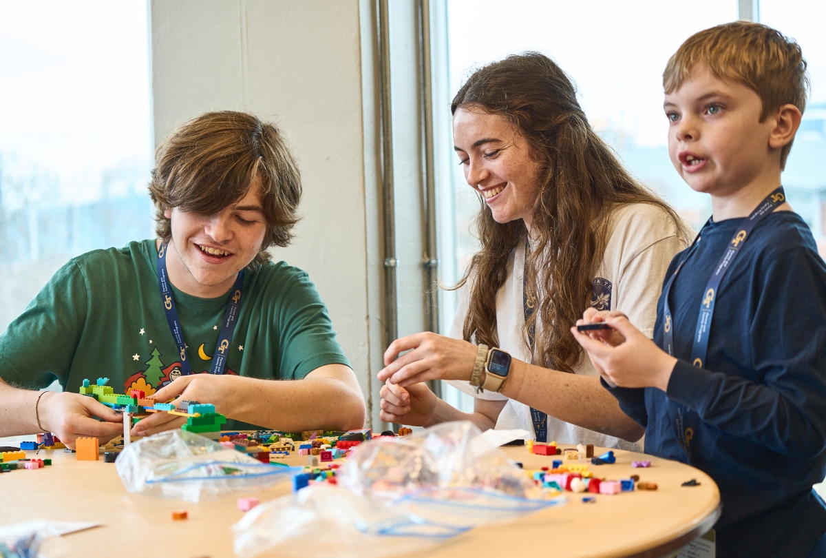 a teenage boy, young adult woman, and young boy sit at a table building with Lego blocks