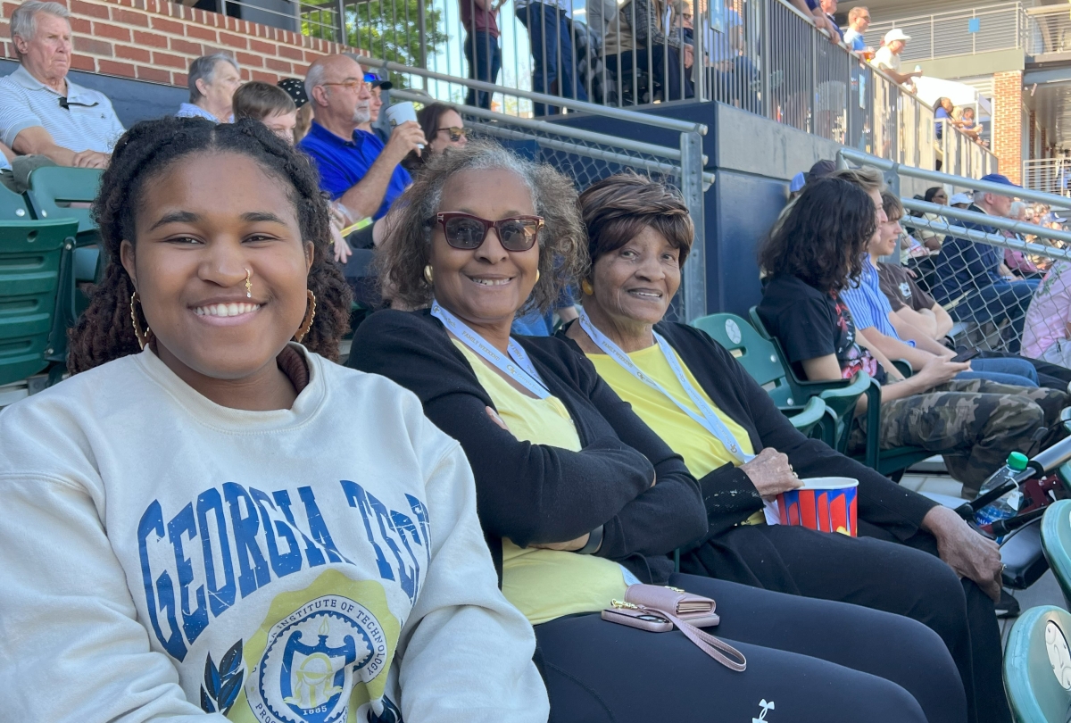 three generations of women sit in sports stadium, smiling at the camera